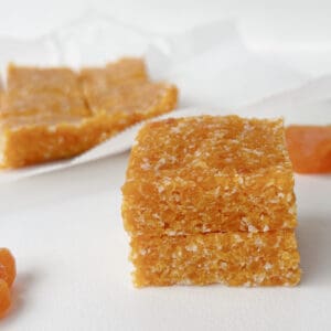 Two pieces of Apricot and Coconut Slice stacked on top of each other. In the background are dried apricots.