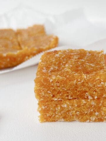 Two pieces of Apricot and Coconut Slice stacked on top of each other. In the background are dried apricots.