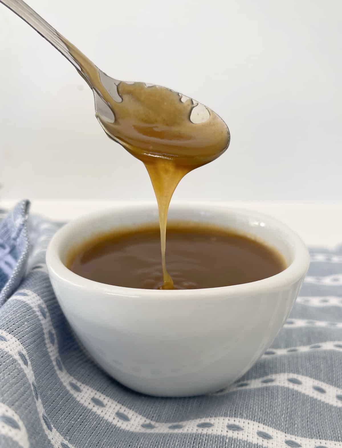 Spoon drizzling salted caramel sauce into a white bowl that is sitting on a blue striped towel.