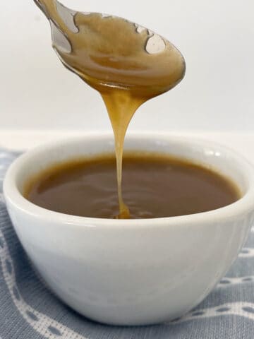 Spoon drizzling salted caramel sauce into a white bowl that is sitting on a blue striped towel.