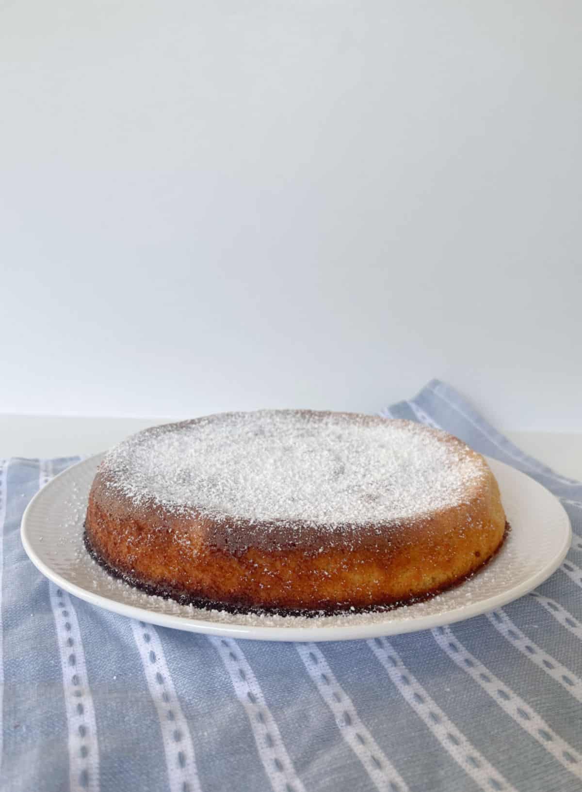 Side view of Whole Orange Cake dusted with icing sugar sitting on a white plate.