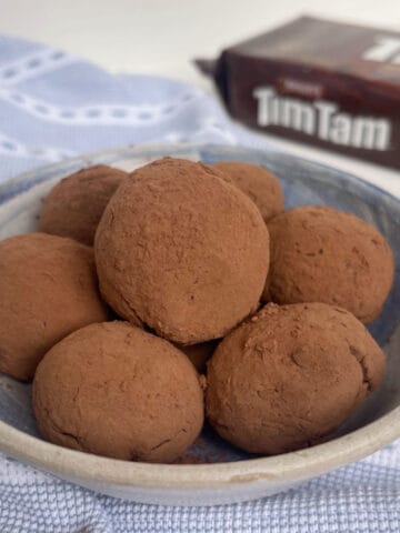 Tim Tam Balls sitting in a blue and white bowl that is on a blue and white striped tea towel. There is a pack of Tim Tams in the background.