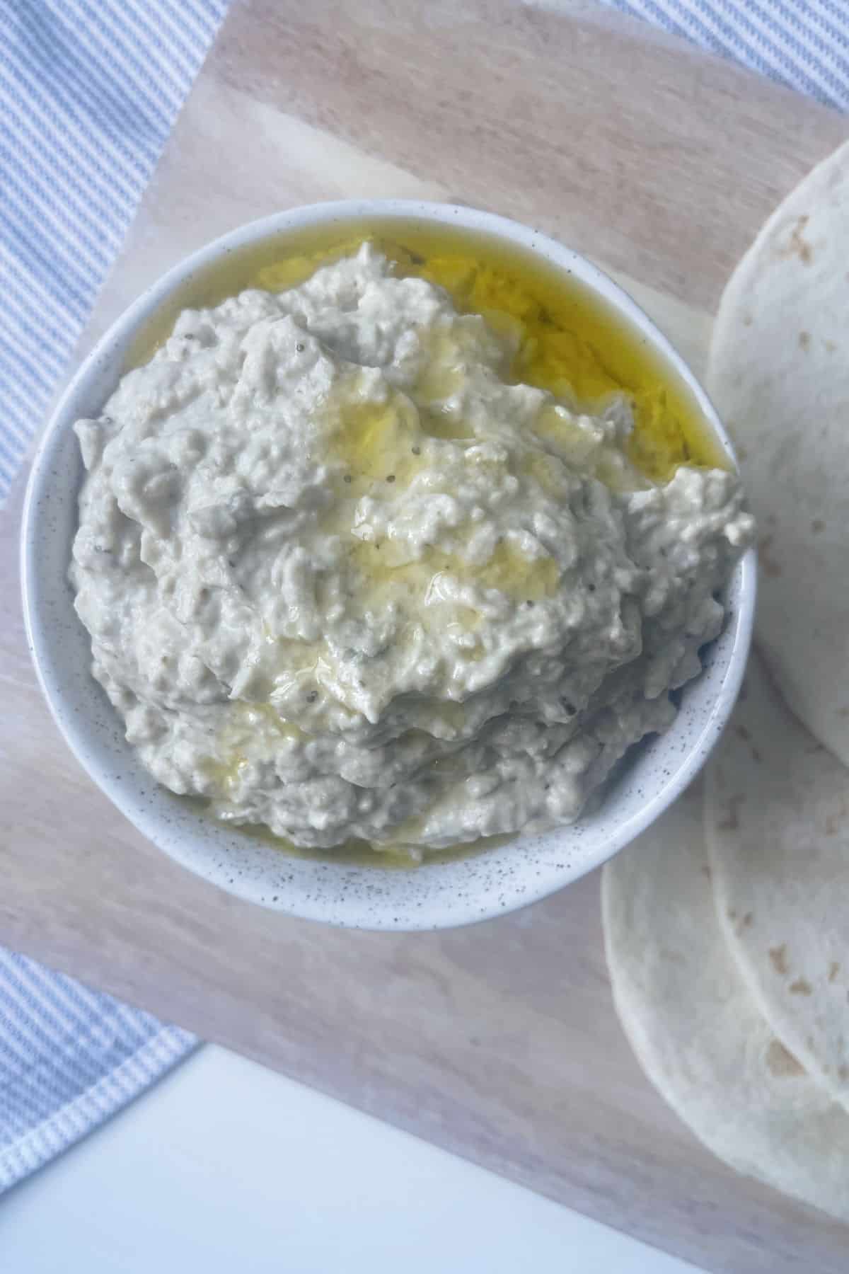 Baba Ghanoush drizzled with olive oil in a white speckled bowl sitting on chopping board.
