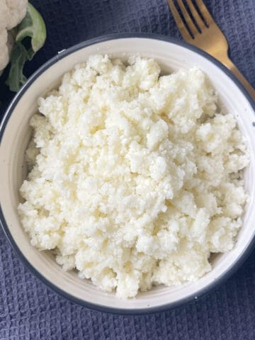 Overhead view of A bowl of Cauliflower Rice sitting on a dark grey towel with a cauliflower in background. There is a gold fork next to the bowl.