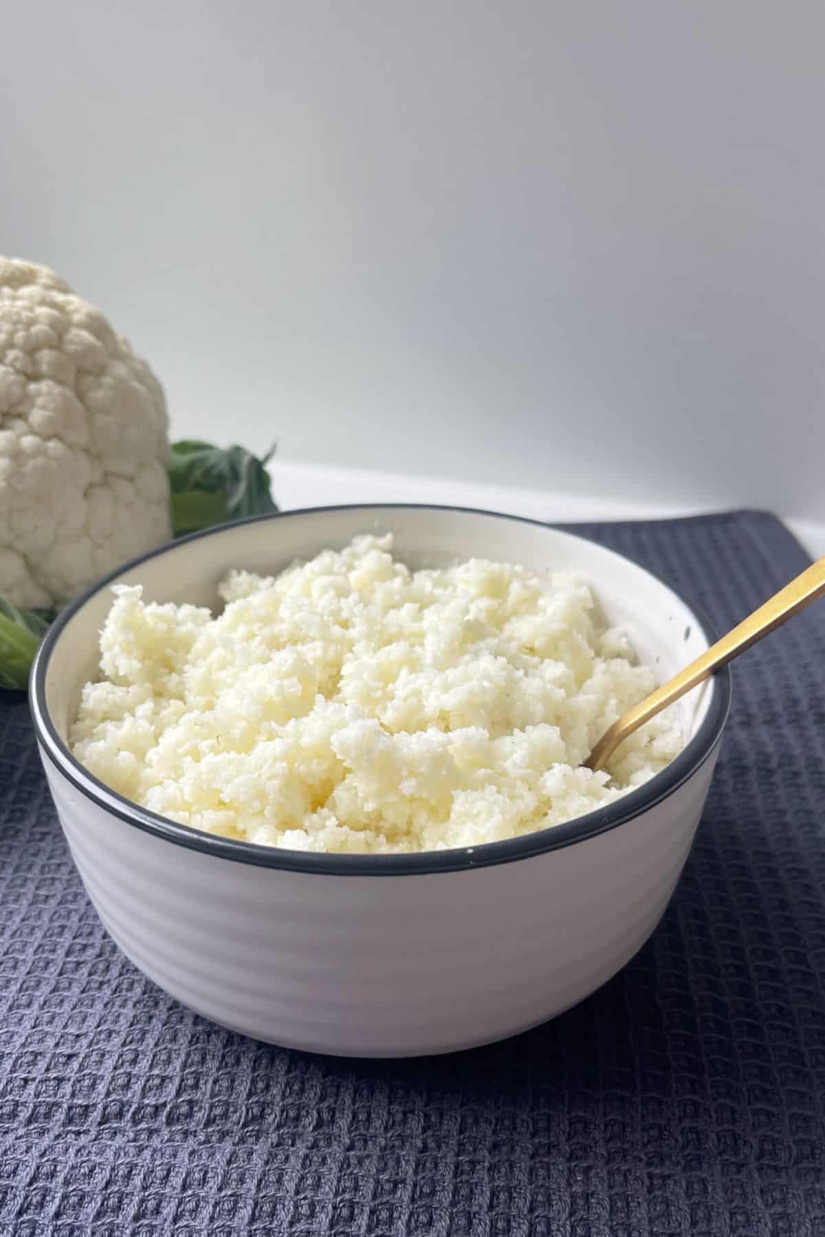 A bowl of Cauliflower Rice sitting on a dark grey towel with a cauliflower in background. There is a gold fork in the bowl.
