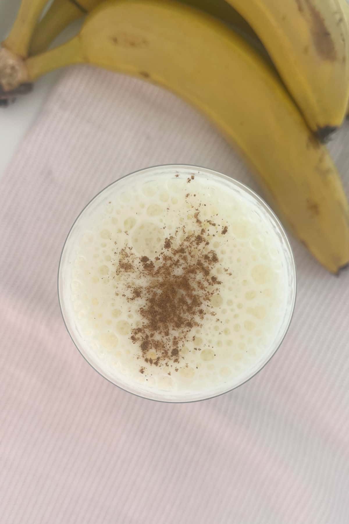 Close up overhead view of a banana smoothie sprinkled with cinnamon. In the background there is a bunch of bananas.