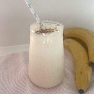 Banana smoothie in a tall glass sitting on a pink towel with a blue striped straw in glass.