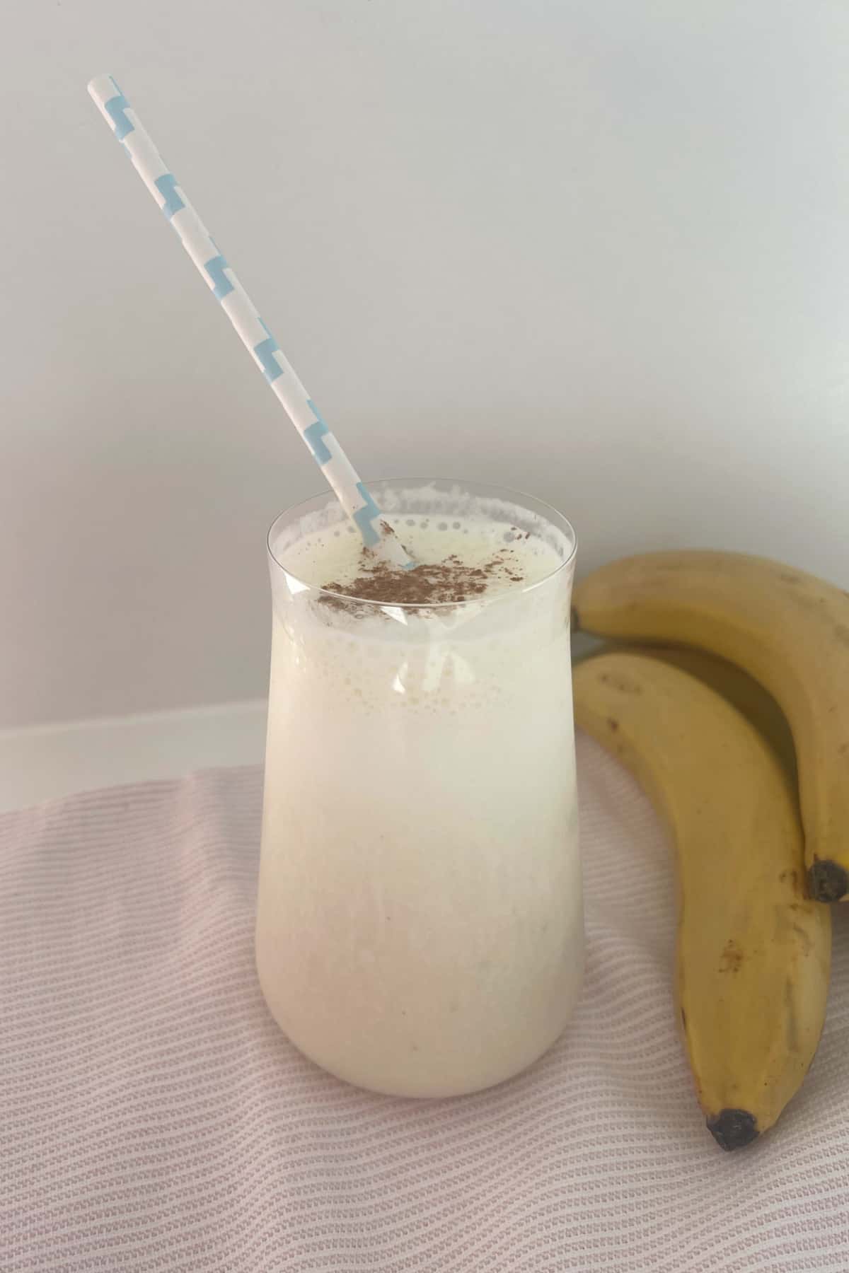 Banana smoothie in a tall glass sitting on a pink towel with a blue striped straw in glass.