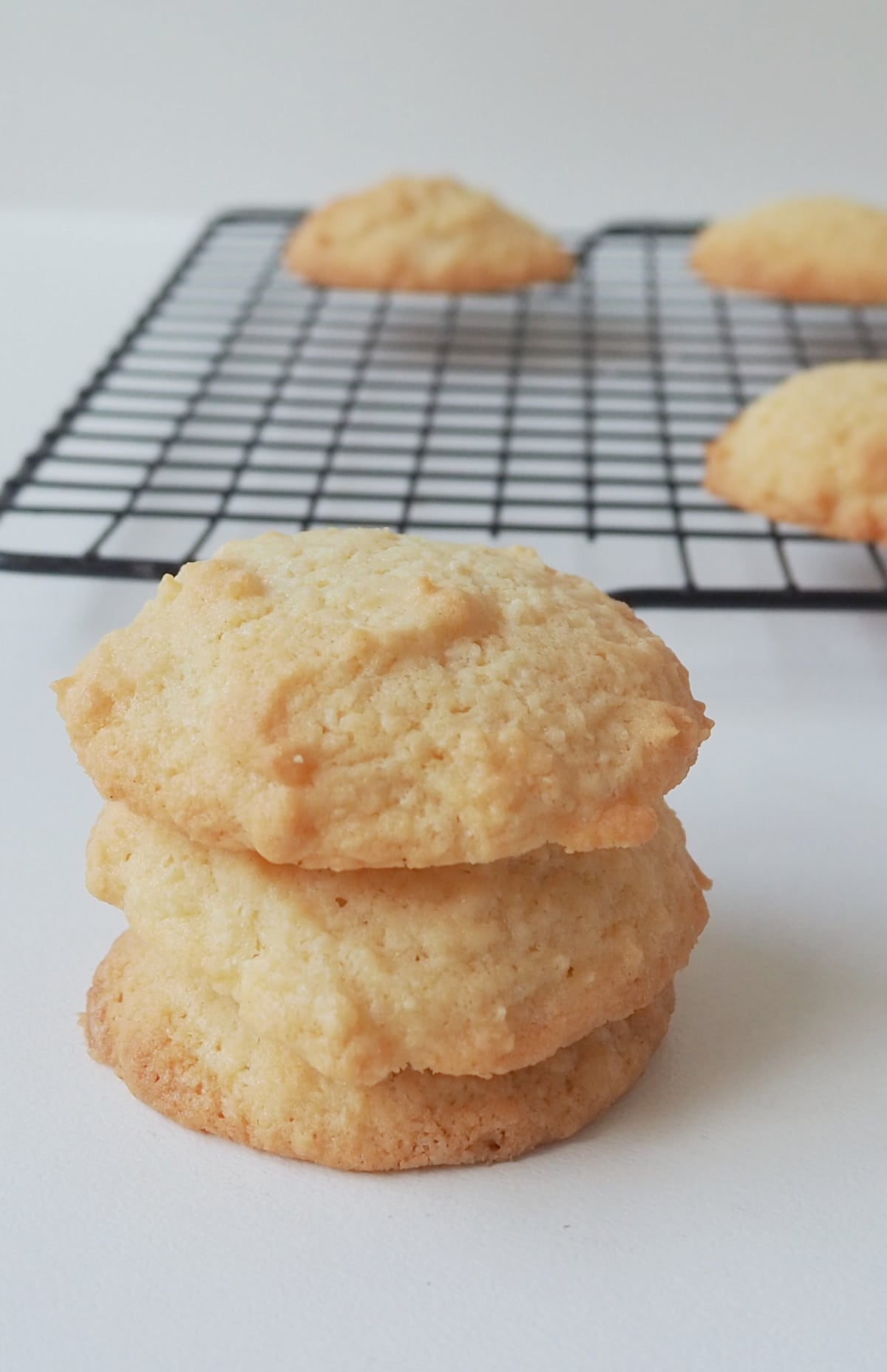 Three coconut biscuits made in a thermomix stacked on top of each other. In the background there is a wire rack with more biscuits on it.