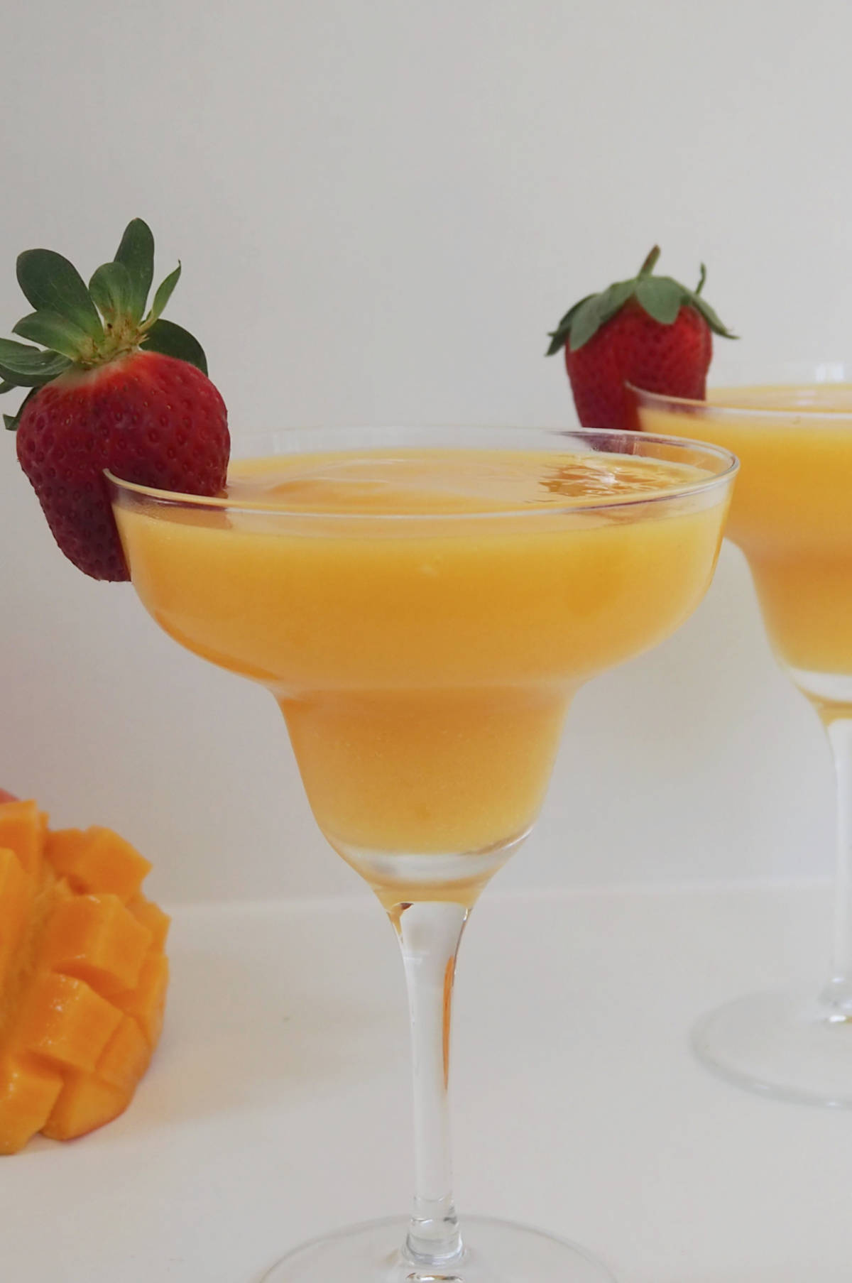 Mango Daiquiri in a cocktail glass and with a strawberry on the rim. In the background is a cut fresh Mango.