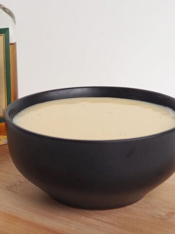 Side view of Brandy Custard in a black bowl sitting on a wooden board. In the background is a bottle of brandy.