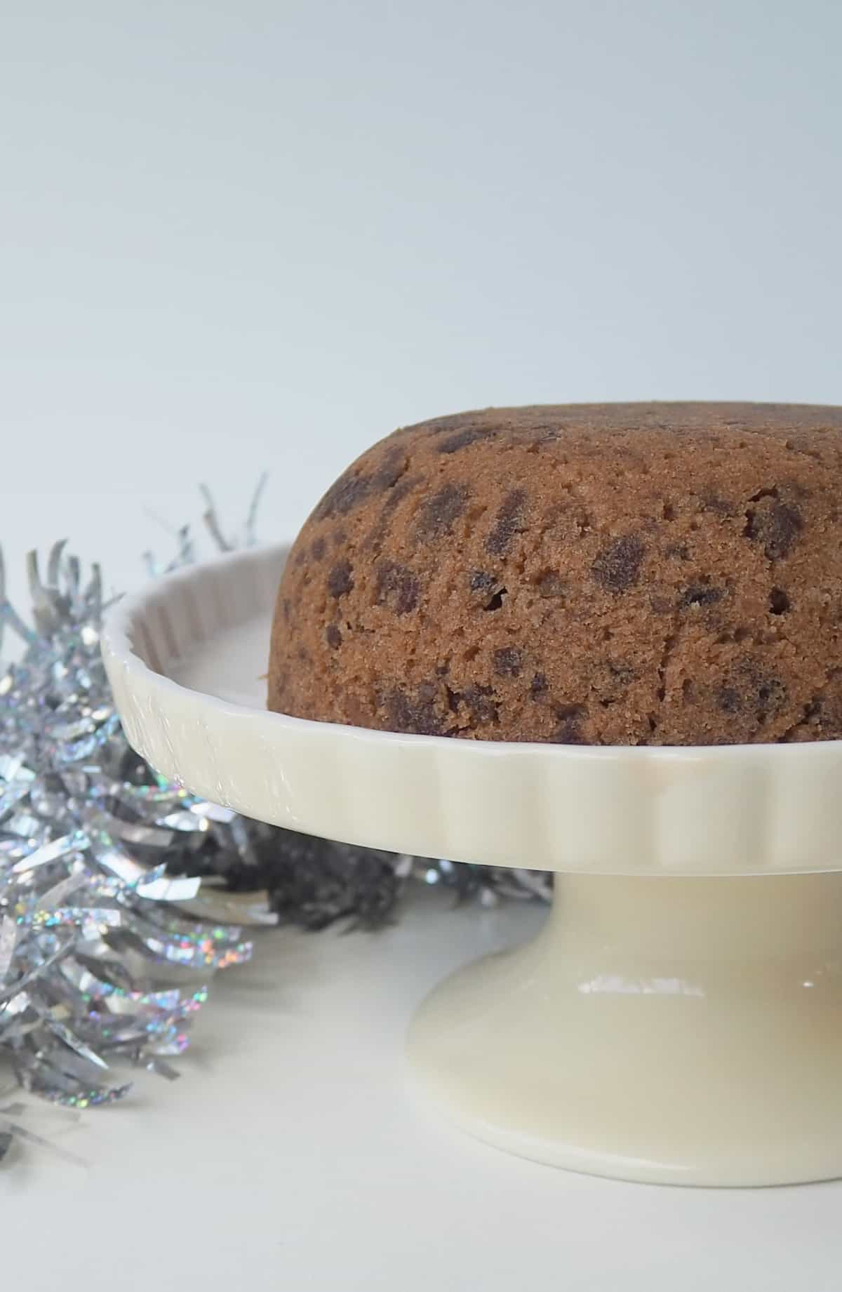 Side view of a Christmas Pudding on a cream cake stand. in the background is silver tinsel.