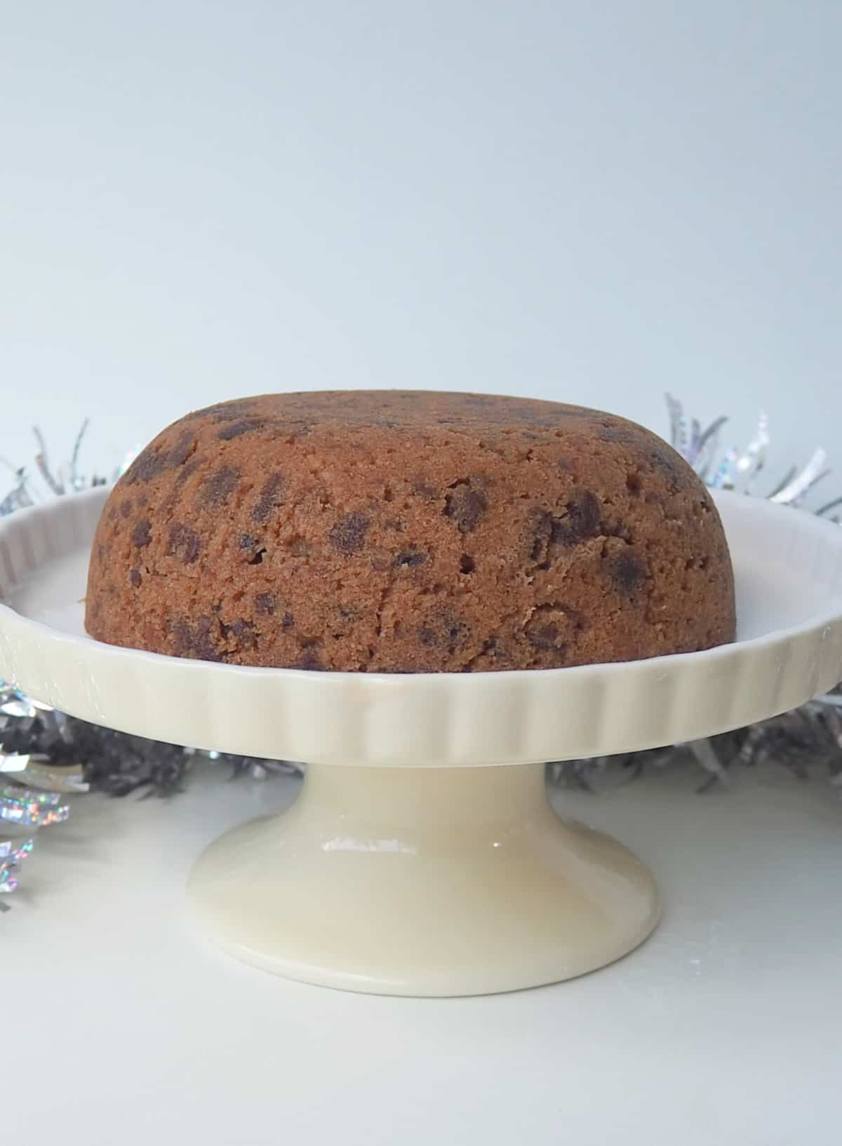 Side view of a Christmas Pudding on a cream cake stand. in the background is silver tinsel.