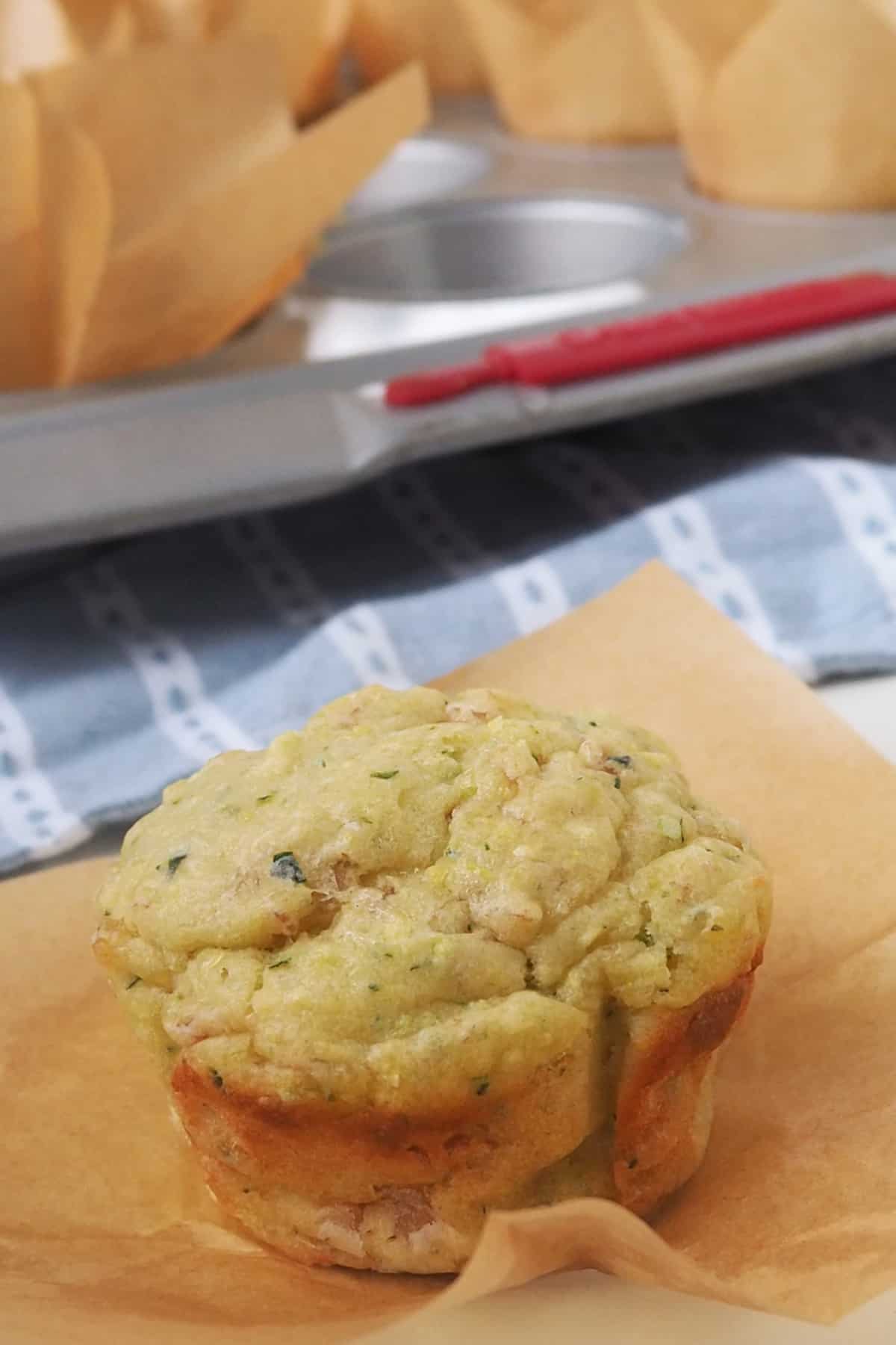 Savoury Muffin in brown paper case on a white surface. In the background is a tray filled with more savoury muffins.