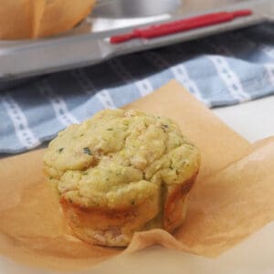 Savoury Muffin in brown paper case on a white surface. In the background is a tray filled with more savoury muffins.