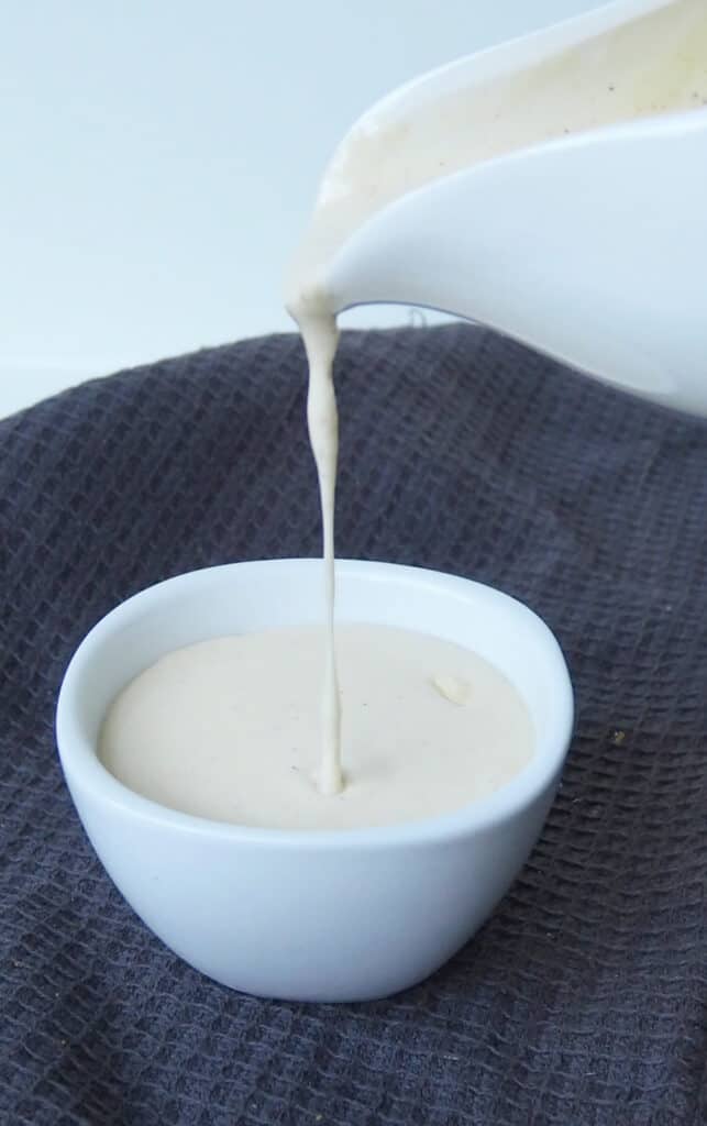 Cheese sauce being poured into a small white bowl that is sitting on top of a dark grey tea towel