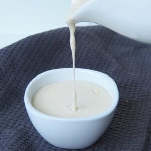 Cheese sauce being poured into a small white bowl that is sitting on top of a dark grey tea towel.