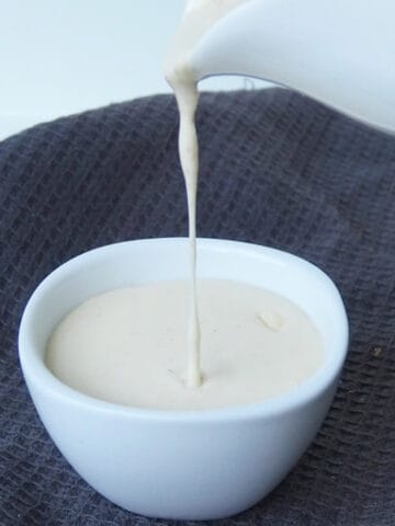Cheese sauce being poured into a small white bowl that is sitting on top of a dark grey tea towel.