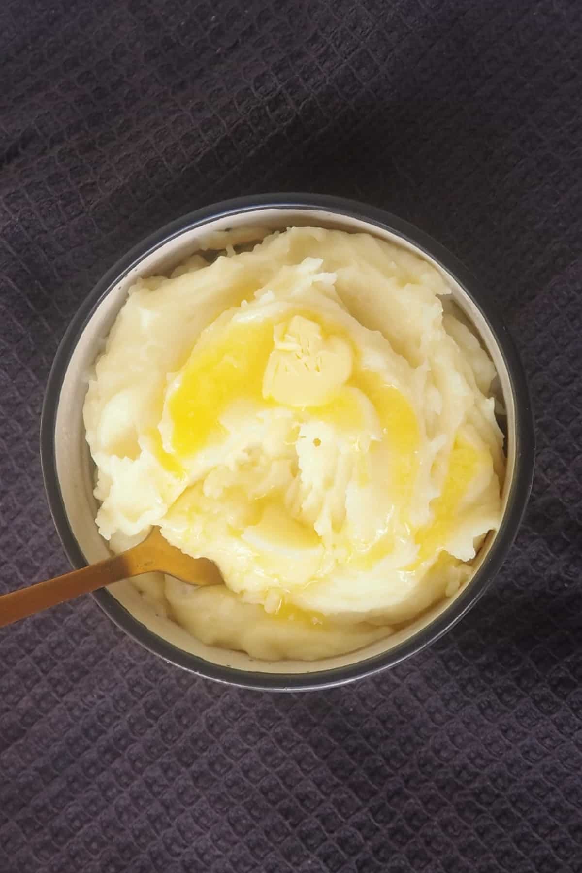 Overhead view of a bowl of mashed potatoes drizzled with butter. There is a gold fork sitting in the potato.