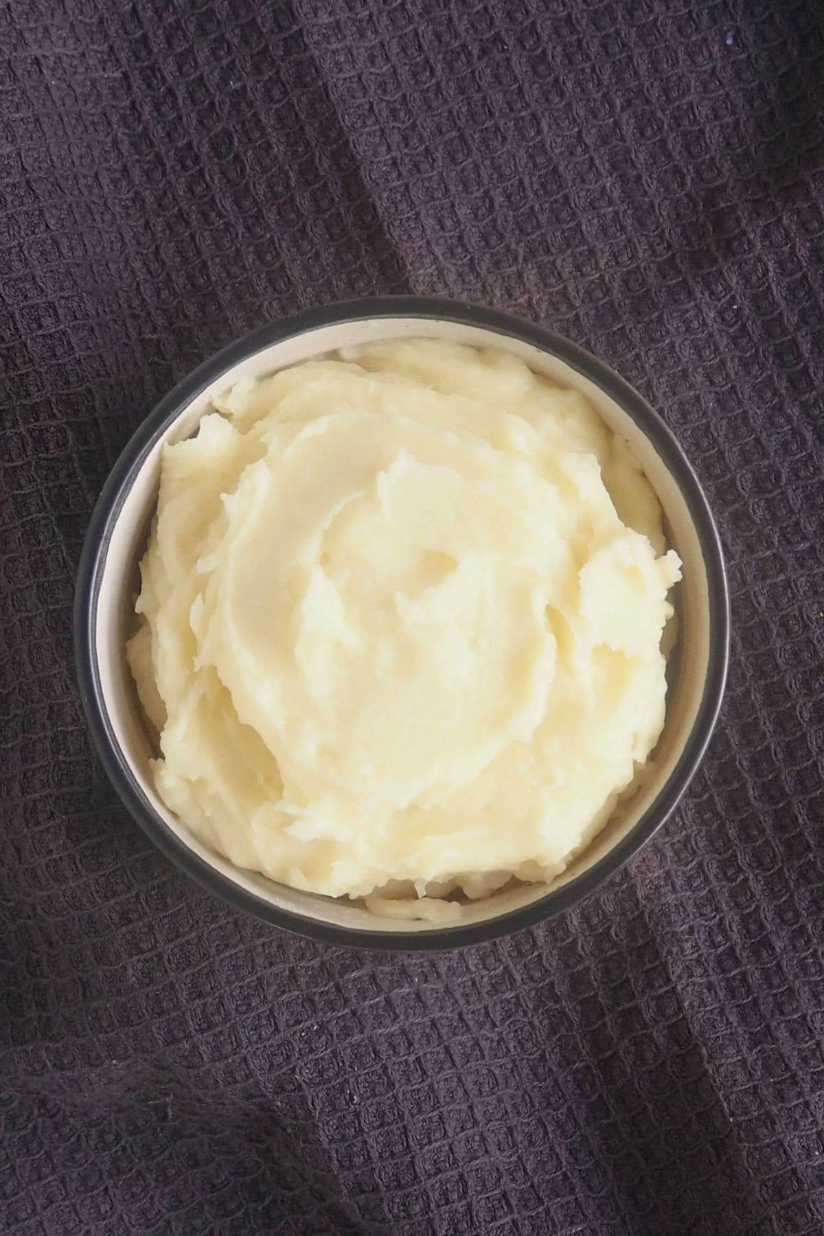 Overhead view of a bowl of mashed potatoes. The bowl is sitting on a dark grey towel.