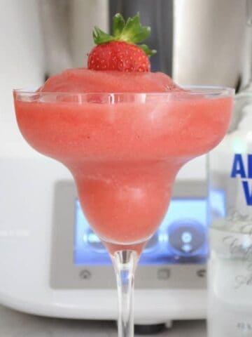 A slushy strawberry drink served in a stemmed cocktail glass with a strawberry on top, and a bottle of vodka nearby.