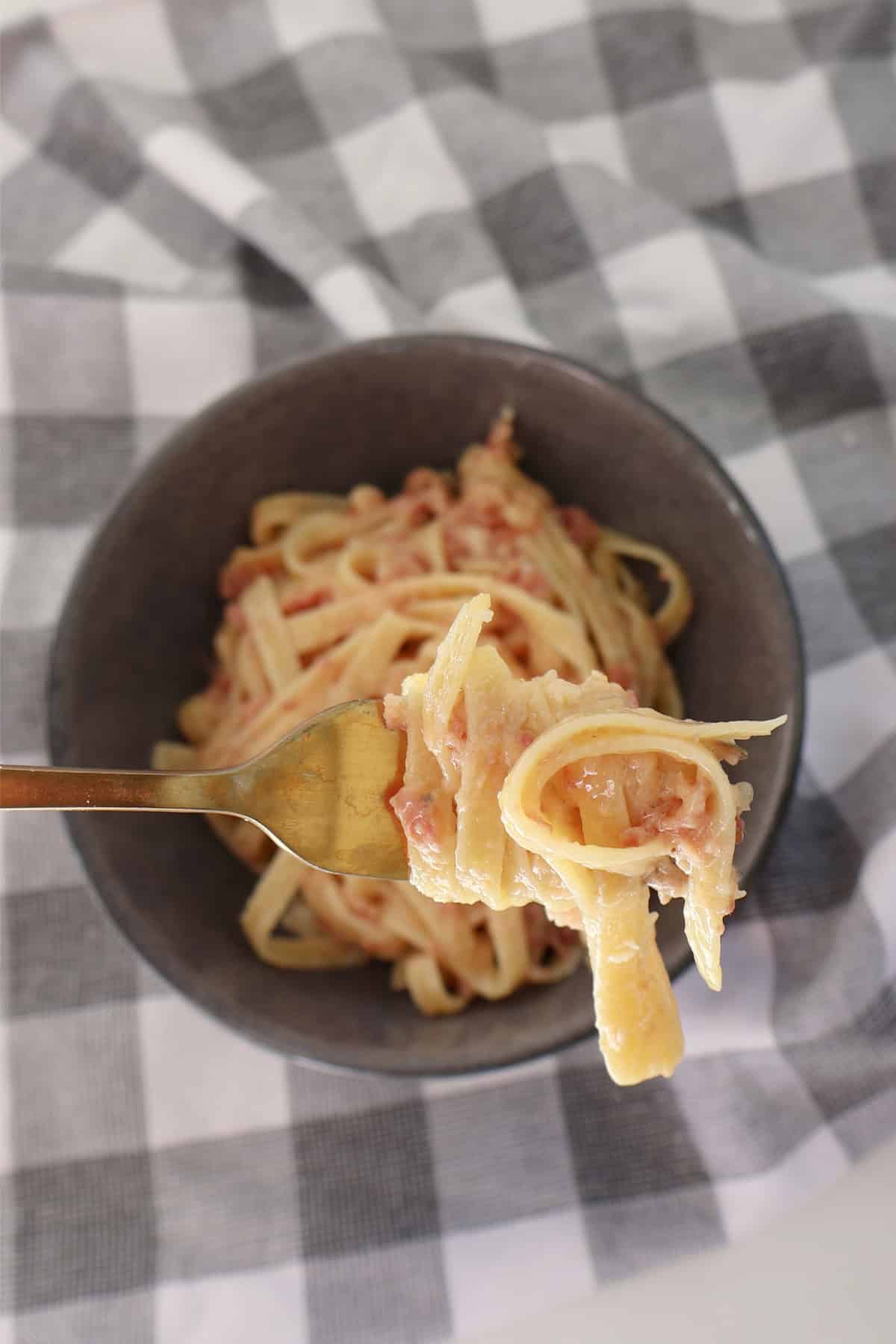 Overhead view of carbonara in a grey bowl. Over the top is a scoop of pasta being held up by a gold fork.